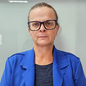 Ana, secretária executiva da Radar DV, usando blazer azul e óculos, em frente a uma parede de vidro.