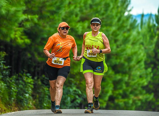 Atleta com deficiência visual e sua guia correndo de mãos dadas por estrada cercada de árvores, durante evento da Radar DV; ambas usam uniformes esportivos e números de identificação.