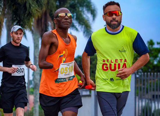 Atleta com deficiência visual corre ao lado de seu guia, usando cordão de ligação, durante prova esportiva do projeto Radar DV; ambos usam óculos escuros e camisetas de identificação.
