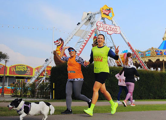 Atleta com deficiência visual corre de mãos dadas com sua guia em evento da Radar DV, ambas sorrindo; ao fundo, parque de diversões com brinquedos e barracas, e um cachorro acompanha a corrida.