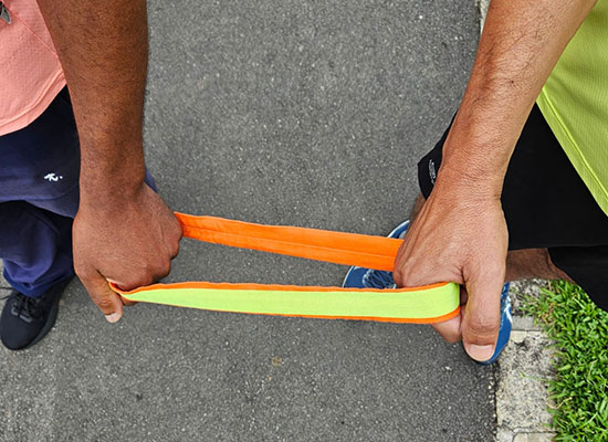 Mãos de um atleta guia e de um atleta com deficiência visual segurando uma corda de conexão laranja e amarela, simbolizando parceria e inclusão durante corrida.