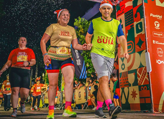 Atleta com deficiência visual da Radar DV cruza a linha de chegada sorrindo, de mãos dadas com seu guia voluntário, durante a Corrida Noite Feliz, em Curitiba. Ambos usam gorros de Natal e transmitem alegria, superação e espírito de inclusão em um ambiente festivo e iluminado.