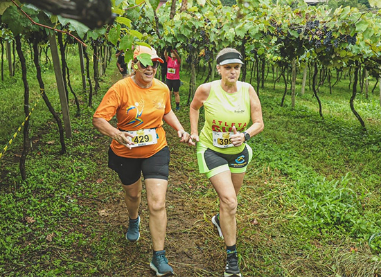 Dupla formada por guia voluntária e atleta com deficiência visual corre por entre parreirais em trilha rural, promovendo a inclusão esportiva com a Radar DV.