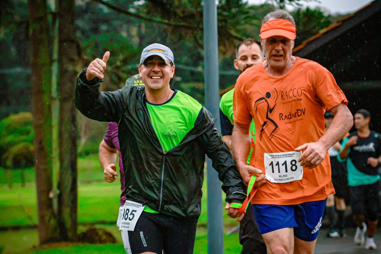 Grupo de corredores da Radar DV participando de corrida inclusiva; pessoas com deficiência visual usam camisetas laranja e são acompanhadas por atletas guias com coletes amarelos. Todos estão fantasiados com saias de bolinha azul e orelhas da Minnie, celebrando o espírito alegre e inclusivo do evento.