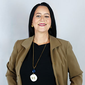 Sandra, tesoureira da Radar DV, sorrindo, usando blazer marrom claro, vestido preto e colar longo com pingentes. Foto em fundo neutro.